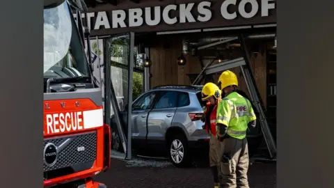 Two fire fighters in yellow helmets and fire fighting overalls stand together while a large silver car is positioned behind them. The car is inside a Starbucks coffee shop and there is glass all around it with a door or window hanging over the car. 