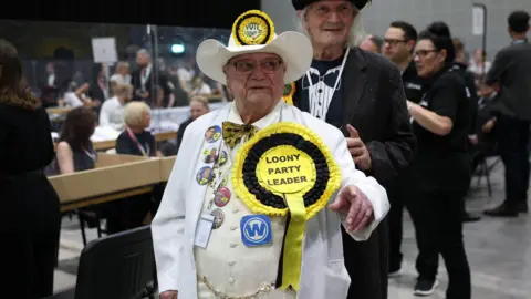 EPA Howling Laud Hope standing in the hall of an election count wears a white suit with a giant yellow rosette which reads "Loony Party Leader". He also has a white cowboy hat with a smaller yellow rosette on it.
