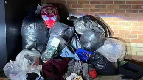 A large pile of bags containing clothes have been piled next to a black collection bin. The bags are being lit by car headlights.