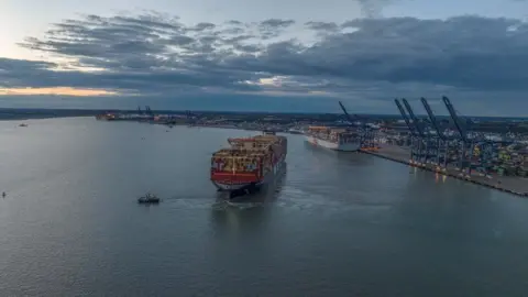 A large container ship and a tug are seen leaving a port. Another large ship is moored at the dock behind