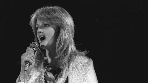 Getty Images Bonnie Tyler is seen in a black and white photograph performing on 15 April 1978. She is singing into a microphone and has feathered shoulder-length hair and is wearing a light-coloured sleeveless top against a dark background.