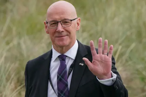 PA Media John Swinney, who wears a dark suit with a tartan tie, arrives on a golf course.