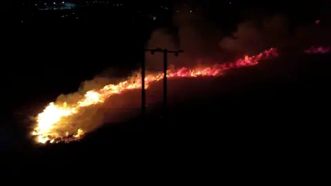 Picture of an orange glowing wall of grass fire ripping across a mountain in the darkness. Flames can be seen tearing through grass, with clouds of smoke rising behind it. A Pylon can be seen on the foreground