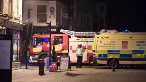 Matt Cardy/Getty Images A red and yellow fire truck parked next to a yellow ambulance on a dimly lit road among shops.