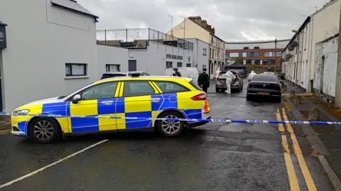 Harford Street in Portadown. In the foreground is a police car, with blue and white tape blocking off the street. Police officers in forensic suits are on the street. 