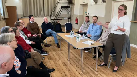 Alex Dunlop/BBC A large group of people sitting in a room hearing a woman talk, who is standing up with her arms out. Three other people are sat at a table close to her with water on it and pieces of paper. Eight other people are sat in a row listening to the woman. The floor is wooden and the walls are cream with cream curtains. 