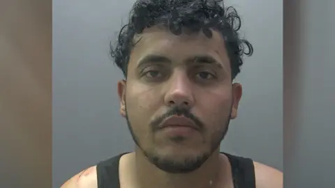 A police custody image of Wail Abduljabbar. He is wearing a black vest and has curly black hair. He is staring straight at the camera and looks moody. He has a scratch on his right shoulder next to his vest.  