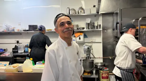 Man wearing chef uniform stood in a kitchen with chefs cooking in the background.