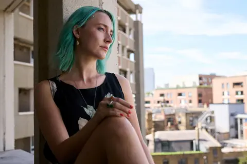 The Outrun/Studio Canal Saoirse, as Rona, with green hair, sitting at the window of a flat in London