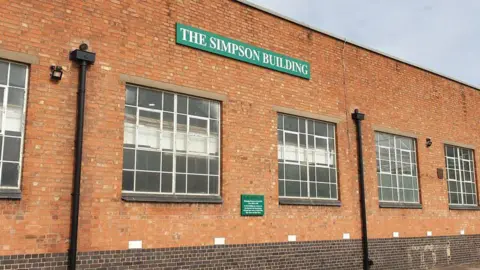 A brick factory with large square windows and a green sign with white lettering that reads The Simpson Building.