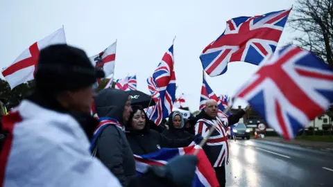 People standing next to a road opposite The Bell Hotel on an overcast day. They are waving flags showing the Union Jack and cross of St George.