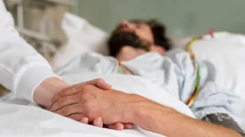A man lying in a bed holding the hand of another person. The man has a beard and is wearing a dark beard.