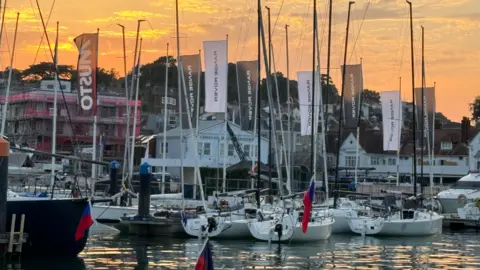 About six small boats with tall masts floating in a marina at sunset ta