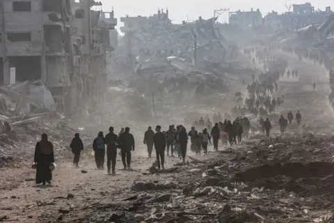 OMAR AL-QATTAA/AFP Scores of displaced Palestinians walk along a road in the Saftawi area of Jabalia, in the Gaza Strip, 19 January 2025.  A crowd of people in silhouette  walk into the distance, amongst the rubble of destroyed buildings.