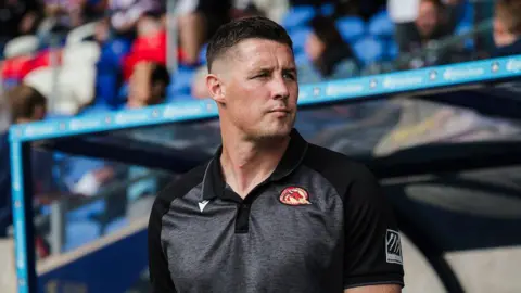 Sam Tomkins standing in the dugout looking over his left shoulder during a game for Catalans Dragons in 2025