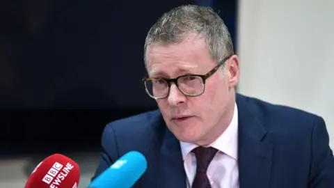Charles McQuilla/Getty Images Kevin Winters, a man with short, greying hair and glasses, leans towards microphones during a press conference in 2024. He is wearing a navy suit, a pink shirt and a dark red tie. 