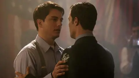 BBC Studios Two men with their arms on each other's arms, gazing in to each other's eyes. One (Gareth David-Lloyd), wearing a navy uniform, has short dark hair and his back to the camera. The other (John Barrowman) also has short dark hair and is wearing a pale blue shirt and suspenders. 