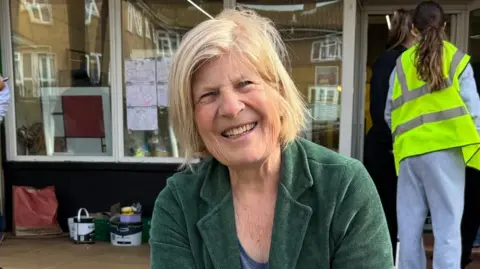 A woman looking at the camera and smiling wearing a green jacket. In the background is a glass shop front and a person in grey jogging bottoms and hi-viz vest