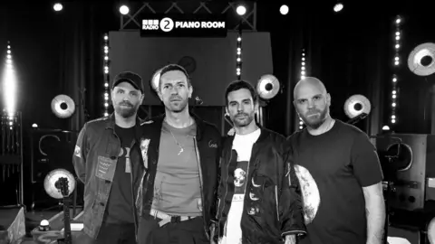 Coldplay are stood in-front of the BBC Piano Room logo. The image is black and white and the mean are stood in a line looking towards the camera. 