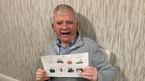 A grey haired man with a grey half zip holds up a sheet with words and pictures on it. He is smiling and happy. 