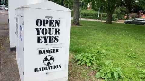 On the side of an electrical box beside a 5G mast are the words "open your eyes" and "danger radiation"