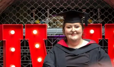 Natasha Dines wears a black mortar board and black robe with red hood. She is sitting in front of large red letters decorated with light bulbs spelling "UWE". 