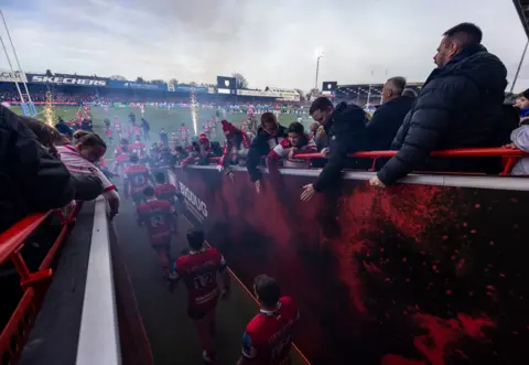 PA Media Fans lean down from the stands to try to high-five Gloucester Rugby players as they walk out of the tunnel and onto the pitch. Pyrotechnics can be seen going off on the field in the background.