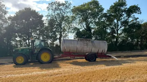 Shropshire Fire and Rescue Service A green and yellow tractor in a field of beige coloured crops is towing a cylindrical water bowser which is pouring water onto the field around a charred area of crops