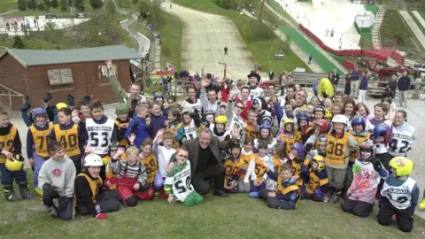 Supplied A large group of children in ski clothes pose with the dry slopes behind them. A man in a brown jacket squats down at the front and waves at the camera.