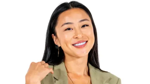 A woman with long, straight, dark hair smiles at the camera. She is Chinese and is wearing a light green top and necklace.