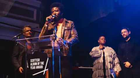 Scottish Album of the Year The singer of the band Young Fathers gives an acceptance speech behind a podium with the words Scottish Album of the Year Award