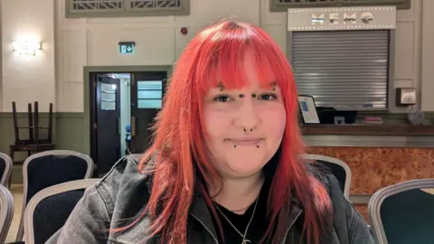 Kasey, a young woman with facial piercings and bright pink hair. She is wearing a black jacket and a black T-shirt