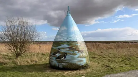 A buoy on grass at a nature reserve. It is painted and features images of birds near a stream. The painting aligns with the horizon. The sky is blue with large grey clouds. 