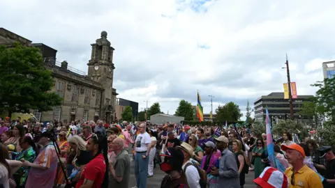 Haydn Brown A large crowd of people standing in Keel Square. They are facing the left-hand side where the stage is and many are holding up flags. There is a large stone building to the left and the sky above is covered in white clouds with a bit of blue peeking through.