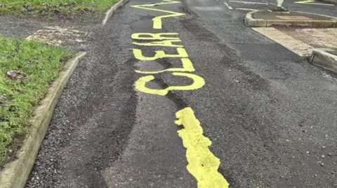 Councillor Andrea Bowes A damaged road surface. Yellow safety markings have been painted onto the road directly over a large pothole. 