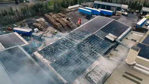 Jack McCausland A large warehouse with smoke coming out of it. There are piles of wood in the background and a number of lorry trailers.