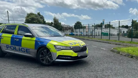 A police car is seen beside a school that has a cordon. 
The school fence is seen outside a large white building.