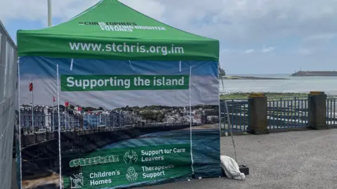 St Christopher’s Isle of Man A gazebo by railings close to the sea. It is green and blue and has writing explaining the work of the St Christopher’s charity.