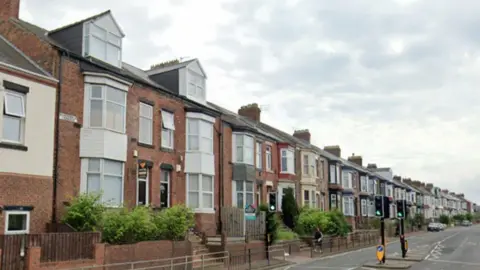 A row of terraced housing sits on the left hand side of the road. A pedestrian crossing with traffic lights sits in the centre of the road. 