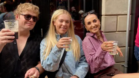 Three people sat, smiling at the camera. The man on the left has blond hair and is wearing a dark shirt and sunglasses. The woman in the middle is wearing a blue denim jacket and has long blonde hair. The woman on the right has dark hair and is wearing a pink and white shirt. They are all holding drinks. 