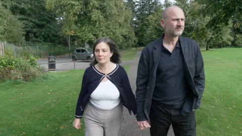 Ros and Mark holding hands arsenic  they locomotion  successful  a park. Mark is wearing a achromatic  garment  and jacket. Ros has a achromatic  apical  connected  with a achromatic  jacket.