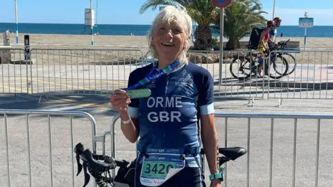 Judy Orme Judy holding her medal which is draped around her neck. Her bike is leaning on a metal fence. She is centre and smiling into the camera. A beach and the sea is in the background.