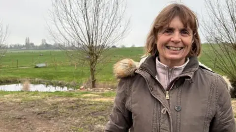 A woman with shoulder length brown hair and a brown zip up winter coat, smiles at the camera. She is standing on the edge of a field, with an automatic animal water drinker in the background. There is a small pond visible behind her.