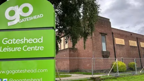 Gateshead Leisure Centre while it was closed. A metal fence has been erected around the brown brick building and its windows have been boarded up. A green sign to the left reads, Gateshead Leisure Centre.