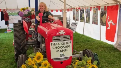 CLAIRE WHITEWAY Claire, sitting on a red tractor bearing the message: Thanks for Choosing Manx on the front, and bunches of sunflowers on either side. The tractor is parked inside a white marquee.