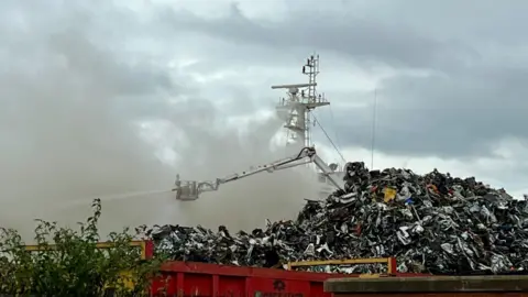 Becki Bowden/BBC A white aerial platform shoots water towards a fire on a ship. A pile of scrap metal is in the foreground of the image.