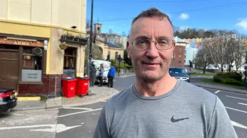 Ciaran Byrne looks at the camera. He has cropped hair and glasses and is wearing a grey Nike t-shirt. The Merchant's Arms frontage is visible in the background.