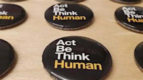 Think Human Several black festival button badges laid on a wooden table. They have the words Act Be Think Human on them.