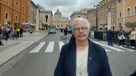 Sister Patricia Murray standing on a street in front of the Vatican. She has short white hair, wearing glasses, a navy jacket, white top, cross brooch, and a long cross necklace.