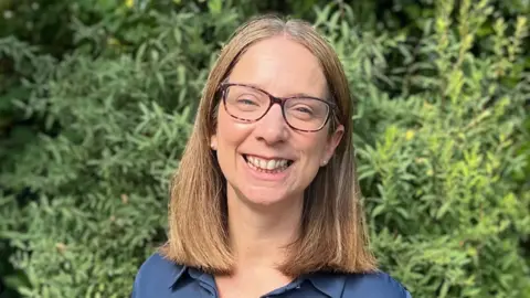 Emma Holland-Lindsay A head-and-shoulders portrait of a woman wearing glasses and a navy-blue shirt, photographed outdoors against a backdrop of lush greenery.
